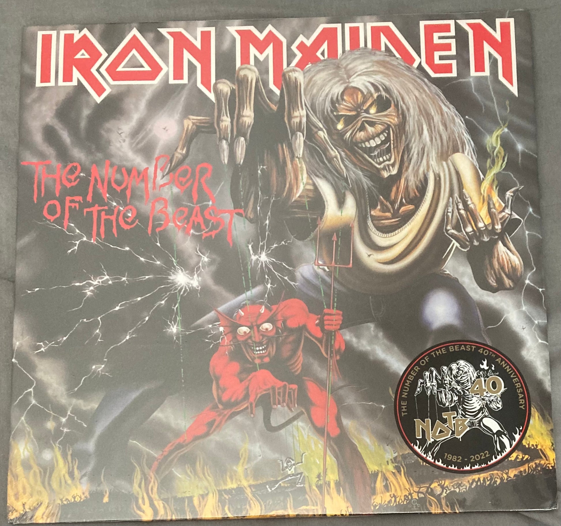 Iron Maiden - The Number of the Beast album cover featuring a stylized illustration of a beast or monster with a red aura, alongside a human figure with a screaming face, all set against a dark, stormy background with lightning. The album title and band name are prominently displayed at the top.